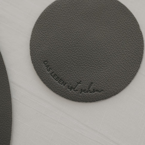 Runder Untersetzer in Dunkelgrau mit Prägung ‚Das Leben ist schön‘ auf weißer Tischdecke.