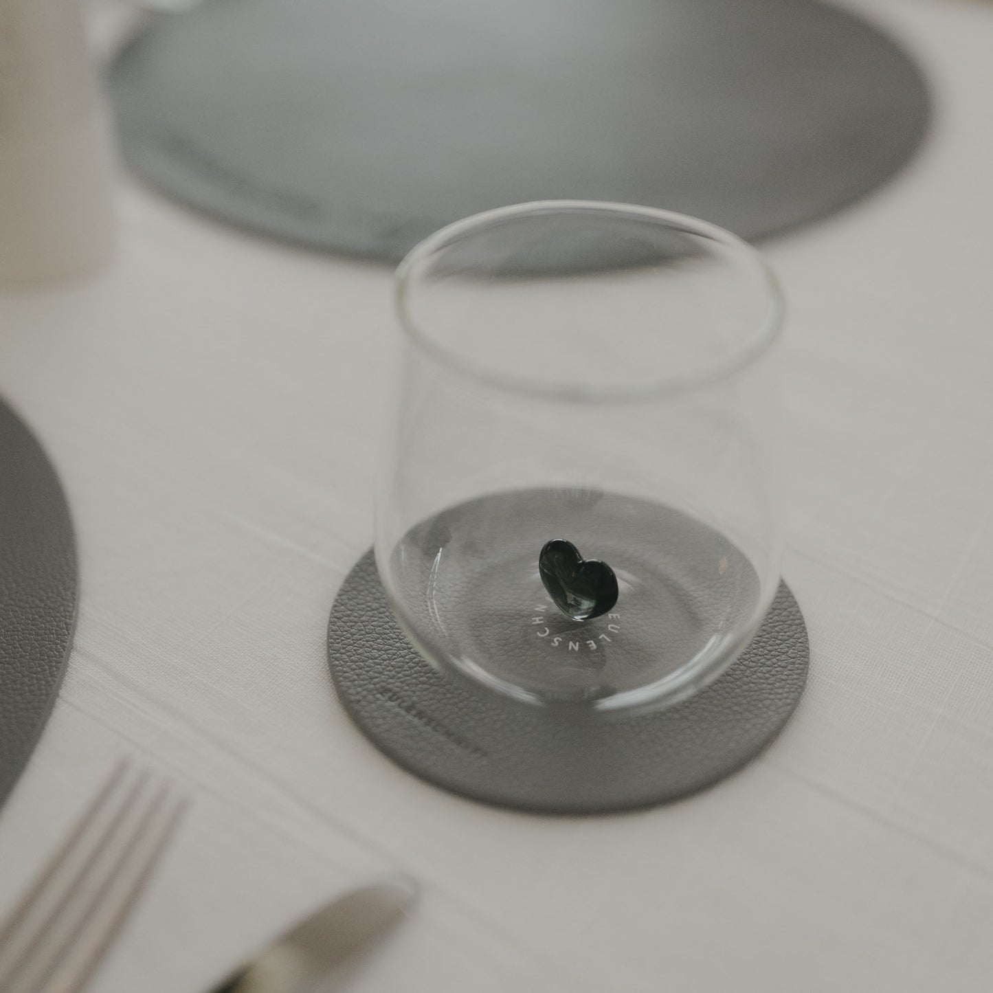 Glas steht auf dunkelgrauem Untersetzer neben Teller und Besteck auf hell gedecktem Tisch.
