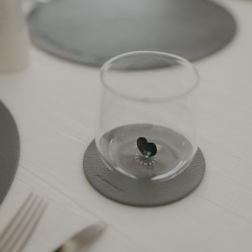 Glas steht auf dunkelgrauem Untersetzer neben Teller und Besteck auf hell gedecktem Tisch.