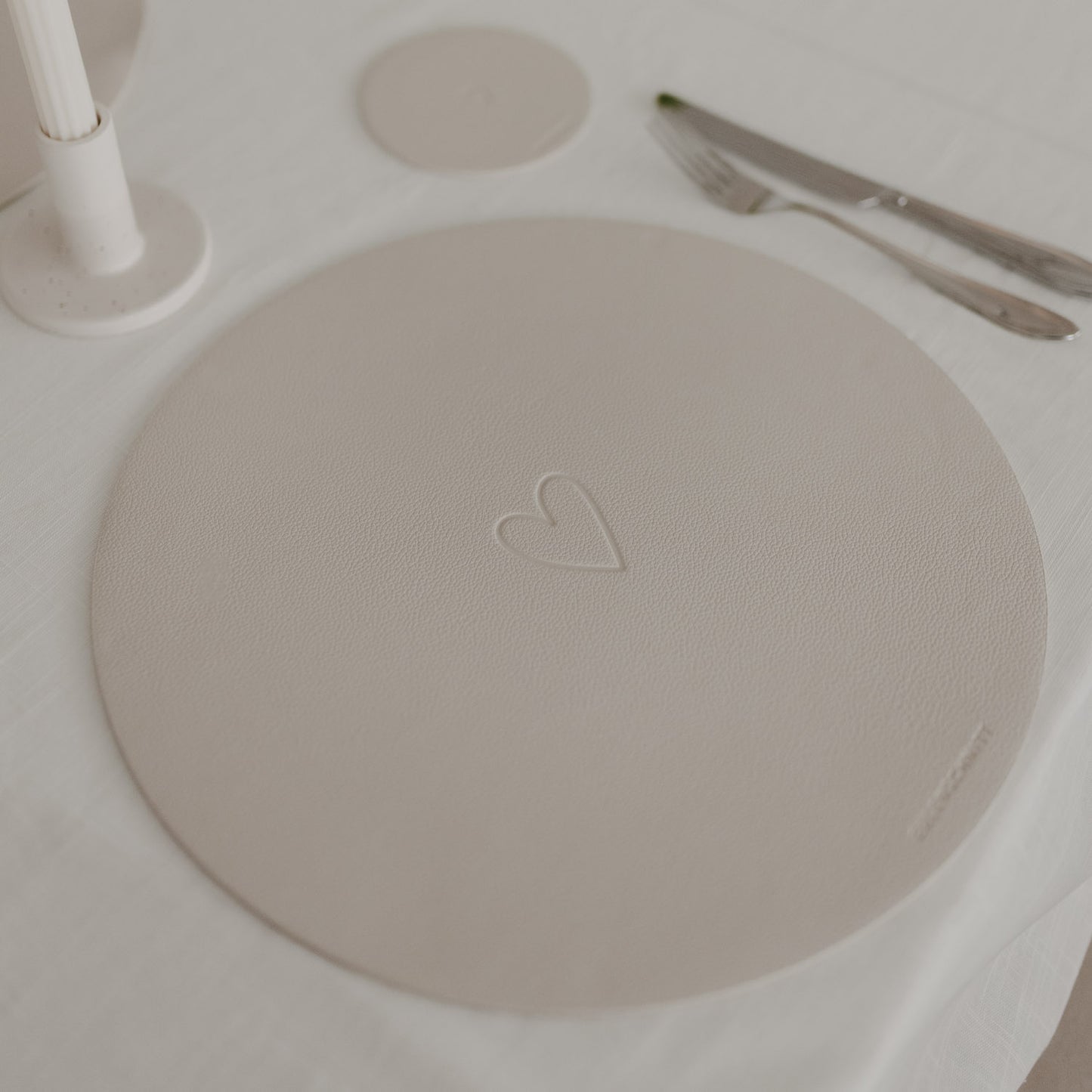 Rundes Platzset mit Herzprägung in Creme, dekoriert auf weißer Tischdecke mit Besteck.