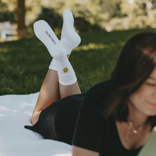 Frau liegt auf Wiese, trägt schwarzes Kleid und weiße Socken mit gelbem Smiley – entspannte Sommerstimmung.