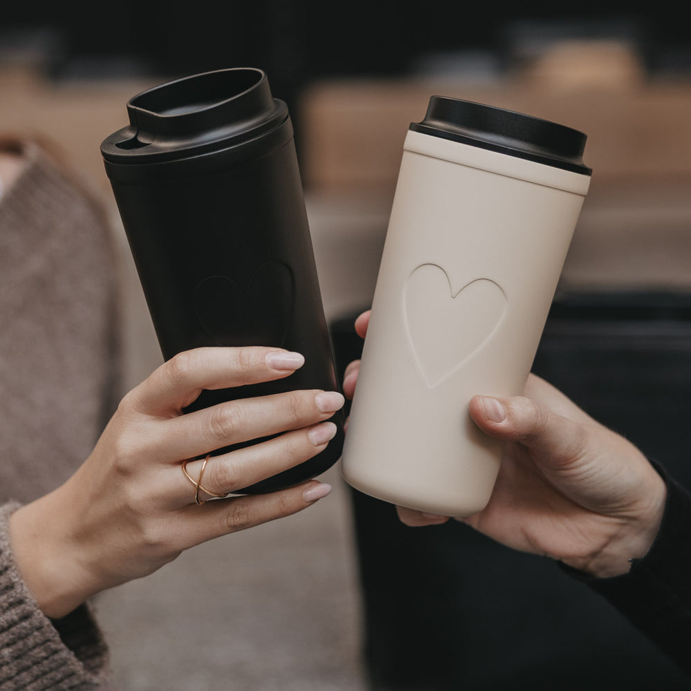 Zwei To-go Becher in Creme und Schwarz mit Herzmotiv, stilvoller Kaffeebecher unterwegs.