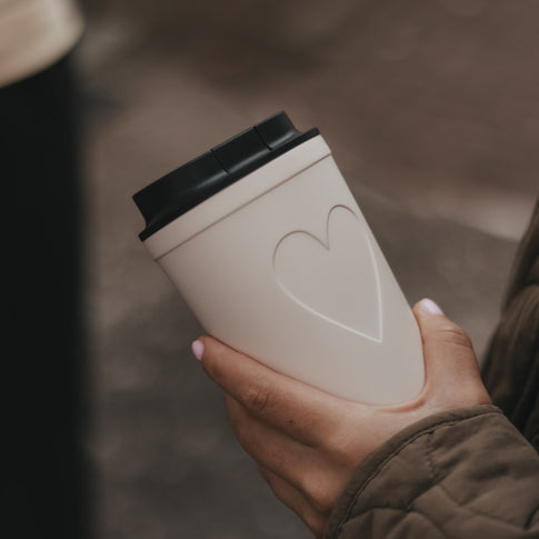 Cremefarbener To-Go-Kaffeebecher mit Herzmotiv und schwarzem Deckel in einer Hand