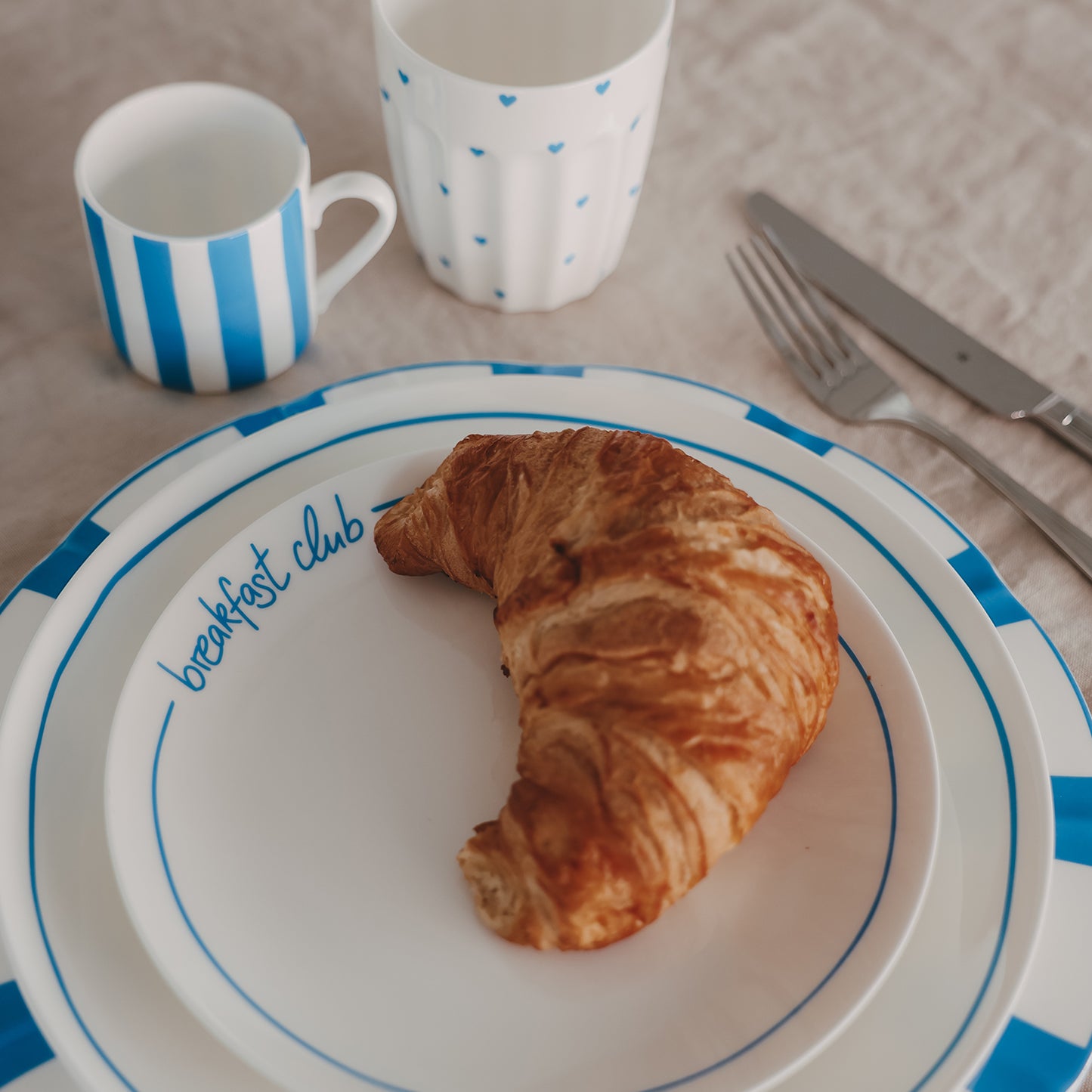 Gestapelte Frühstücksteller mit blauem Streifenrand und „breakfast club“ Schrift, inszeniert mit Croissant und passendem Becher auf Leinentischdecke.