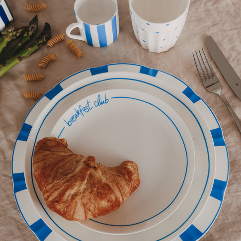 Gestapelte Teller mit blauem Streifenrand im Breakfast-Setting, kombiniert mit Croissant, Tassen, Spargel und Besteck auf natürlicher Leinentischdecke.