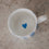 Blick in eine kleine Porzellan-Tasse mit weißem Inneren und einem kleinen blauen Herz am Boden, auf heller Steinfläche.