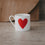 Kleine weiße Tasse mit großem rotem Herz auf der Vorderseite, schlicht inszeniert vor warmem Hintergrund.