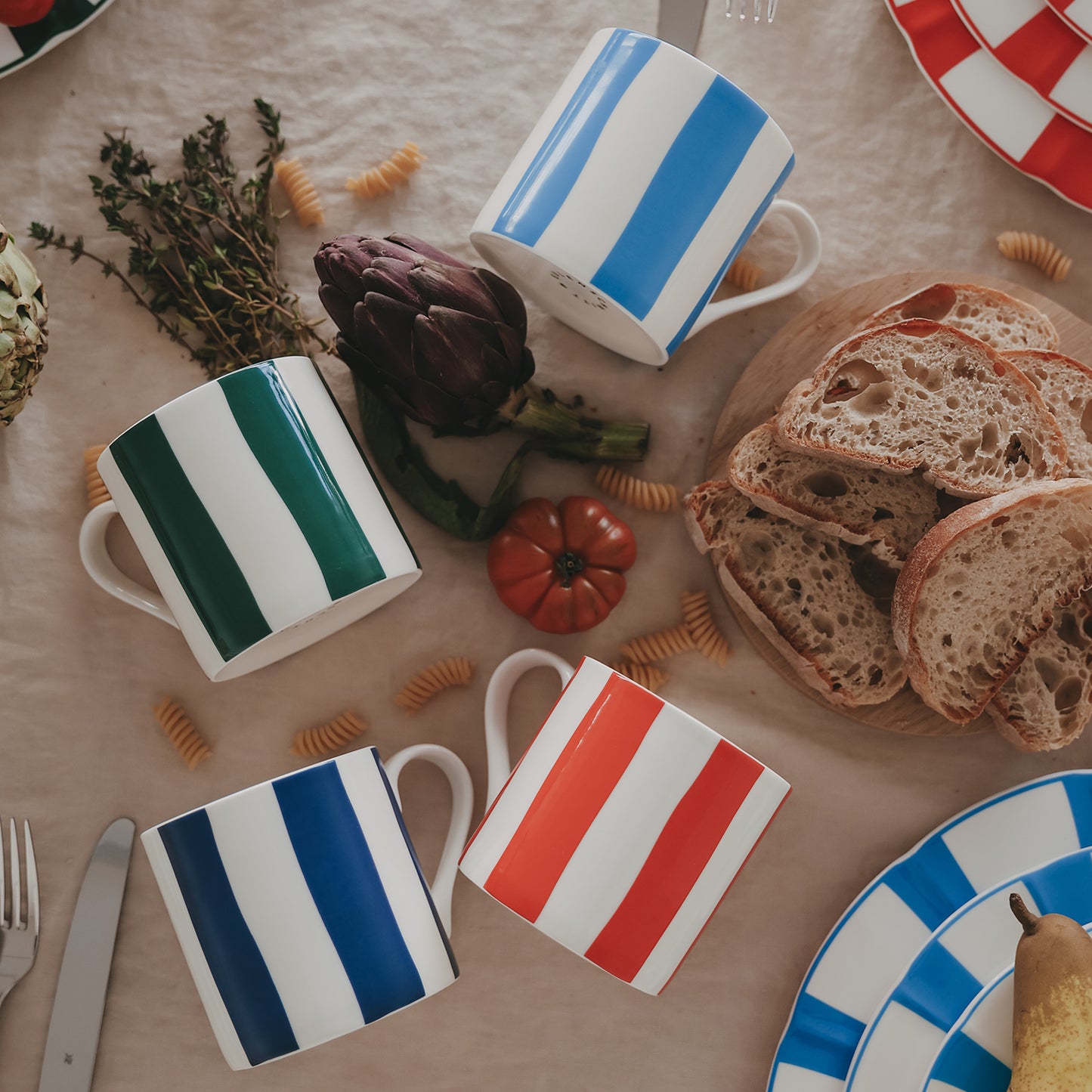 Stillleben mit gestreiften Tassen in verschiedenen Farben, kombiniert mit Brot, Gemüse und Pasta auf einem gedeckten Tisch.