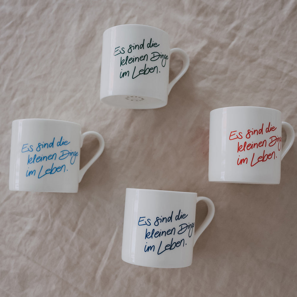 4 weiße Tassen mit Schriftzug „es sind die kleinen dinge im leben“ in verschiedenen Farben auf hellem Untergrund