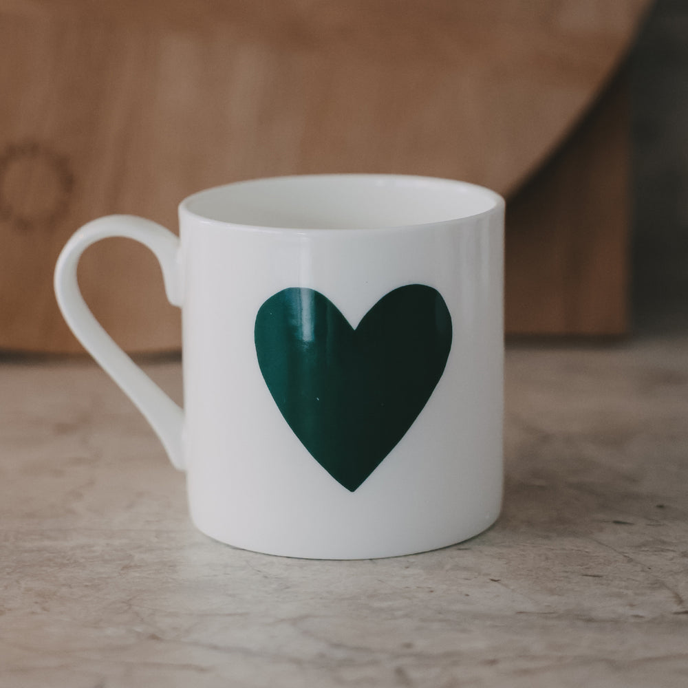 Weiße Tasse mit großem grünem Herzmotiv auf der Außenseite vor schlichtem Hintergrund