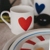 Weiße Tasse mit rotem Herzmotiv auf gedecktem Tisch, im Hintergrund weiteres Geschirr mit blauem Herz