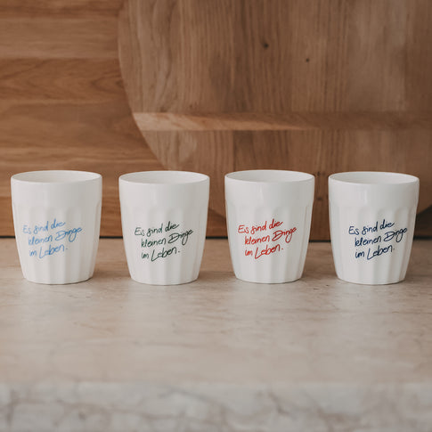 Vier weiße Becher mit Spruch „Es sind die kleinen Dinge im Leben“ in verschiedenen Farben nebeneinander