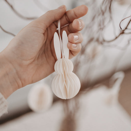 Hand hält beige Papieranhänger in Hasenform vor Osterzweigen.