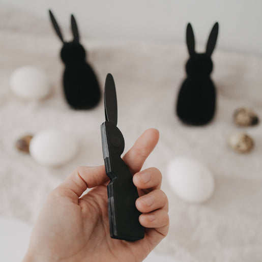 Schwarzer Papierhase in der Hand, weitere schwarze Papierhasen und Ostereier unscharf im Hintergrund.