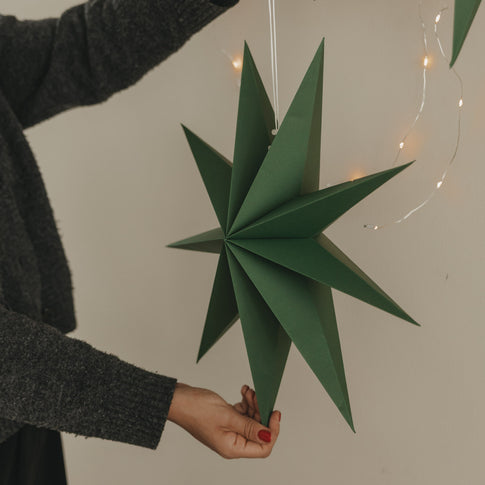 Dunkelgrüner Papierstern mit langen Spitzen und weißer Aufhängung vor warmer Lichterkette