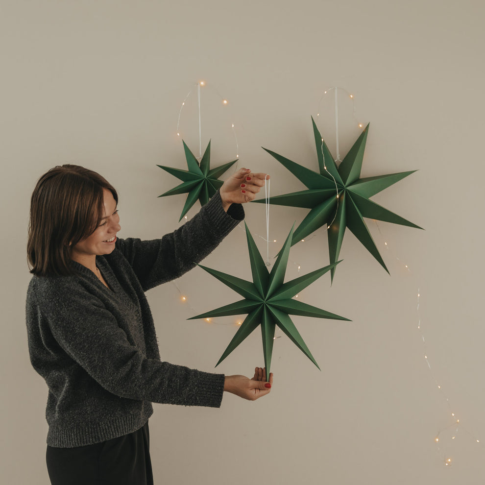 Drei dunkelgrüne Papiersterne mit Lichterkette, einer davon wird dekorativ in der Hand gehalten