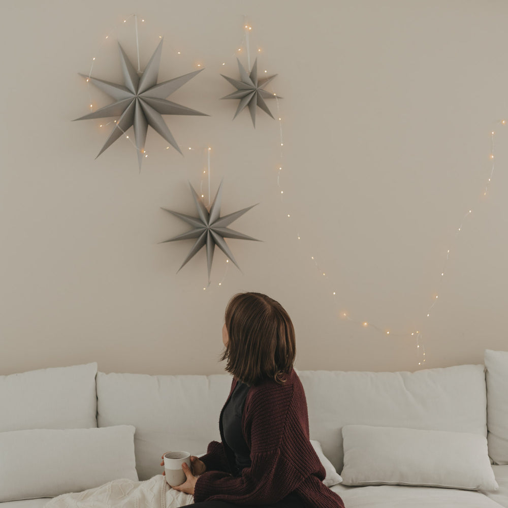 Graue Papiersterne mit warmer Lichterkette als stilvolle Weihnachtsdeko an heller Wand