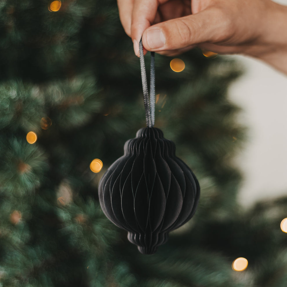 Schwarzer Papieranhänger in Kugelform wird vor einem festlich beleuchteten Weihnachtsbaum gehalten