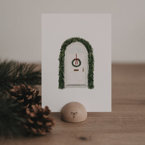 Weihnachtskarte mit weißer Tür, geschmückt mit grünem Türkranz und Tannengirlande