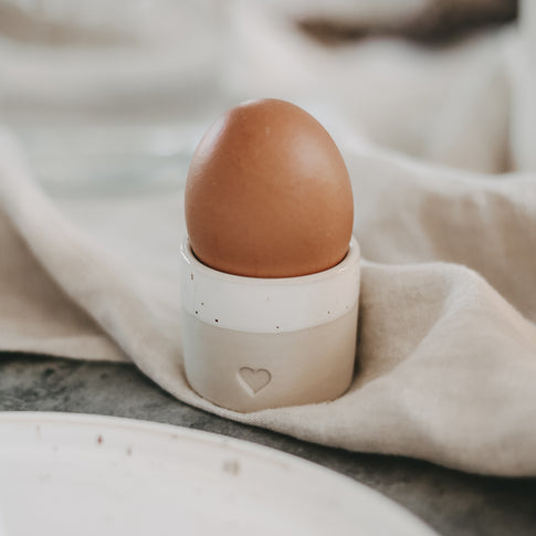 Keramischer Eierbecher in Naturfarben mit Herzprägung und glasiertem Rand, bestückt mit braunem Ei, auf Leinentuch arrangiert.