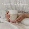 Eine junge Frau sitzt im Bett und hält dabei einen Becher mit der Prägung "Guten Morgen" in ihrer Hand.