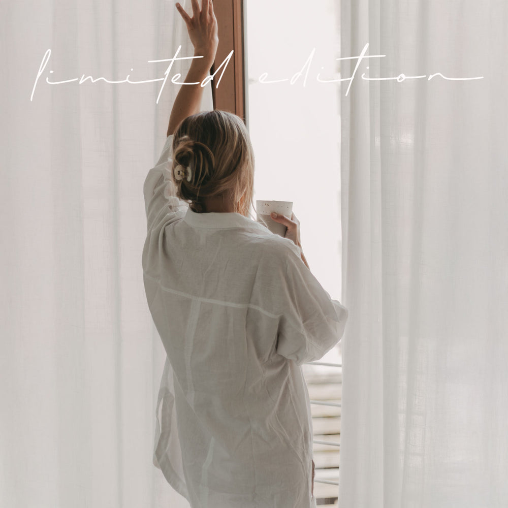 Eine junge Frau mit einer locker gebunden Zopf schaut verträumt aus dem Fenster und schiebt einen weißen Vorhang beiseite. Dabei hält sie einen Becher mit der Prägung "Guten Morgen" in ihrer Hand.