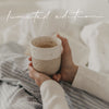 Ein handgetöpferter Becher mit der Prägung "Lieblingsbecher" wird gehalten und die Person sitzt gemütlich in einem hellbau gestreiften Pyjama auf dem Bett.