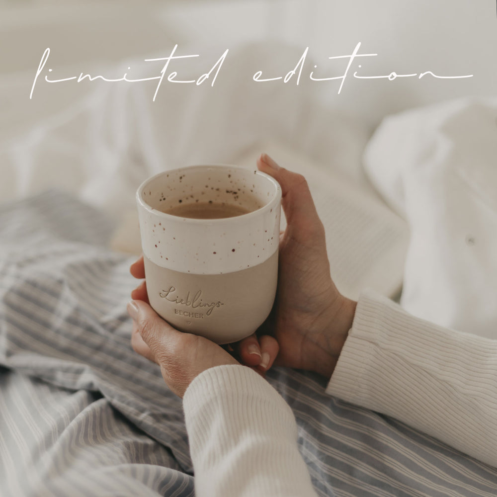 Ein handgetöpferter Becher mit der Prägung "Lieblingsbecher" wird gehalten und die Person sitzt gemütlich in einem hellbau gestreiften Pyjama auf dem Bett.