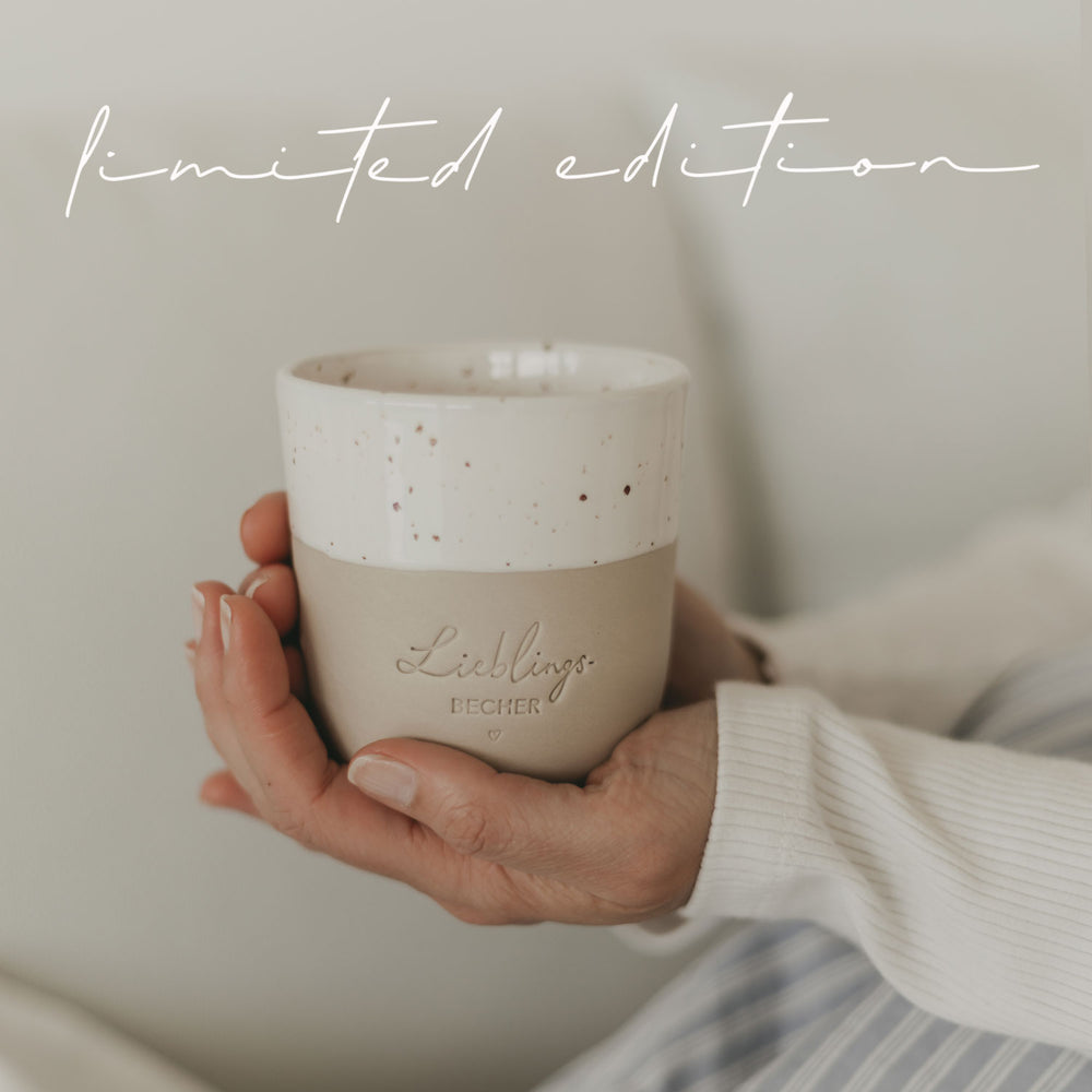 Ein handgetöpferter Becher mit der Prägung "Lieblingsbecher" wird gehalten und die Person sitzt gemütlich in einem hellbau gestreiften Pyjama auf dem Sofa.