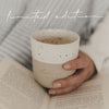 Ein handgetöpferter Becher mit der Prägung "Lieblingsbecher" wird gehalten über einem aufgeschlagenem Buch. Die Person sitzt gemütlich in einem hellbau gestreiften Pyjama auf dem Bett.