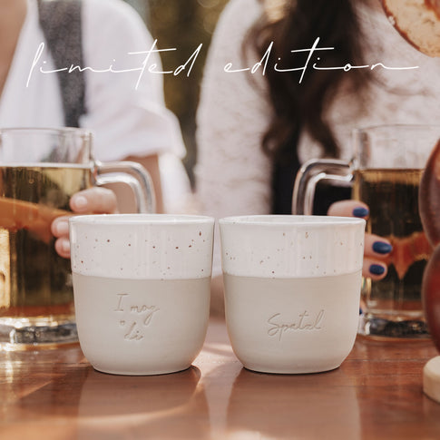 Zwei limitierte Becher „I mog di“ und „Spatzl“ mit Bierkrügen im Hintergrund auf Holztisch im Freien.