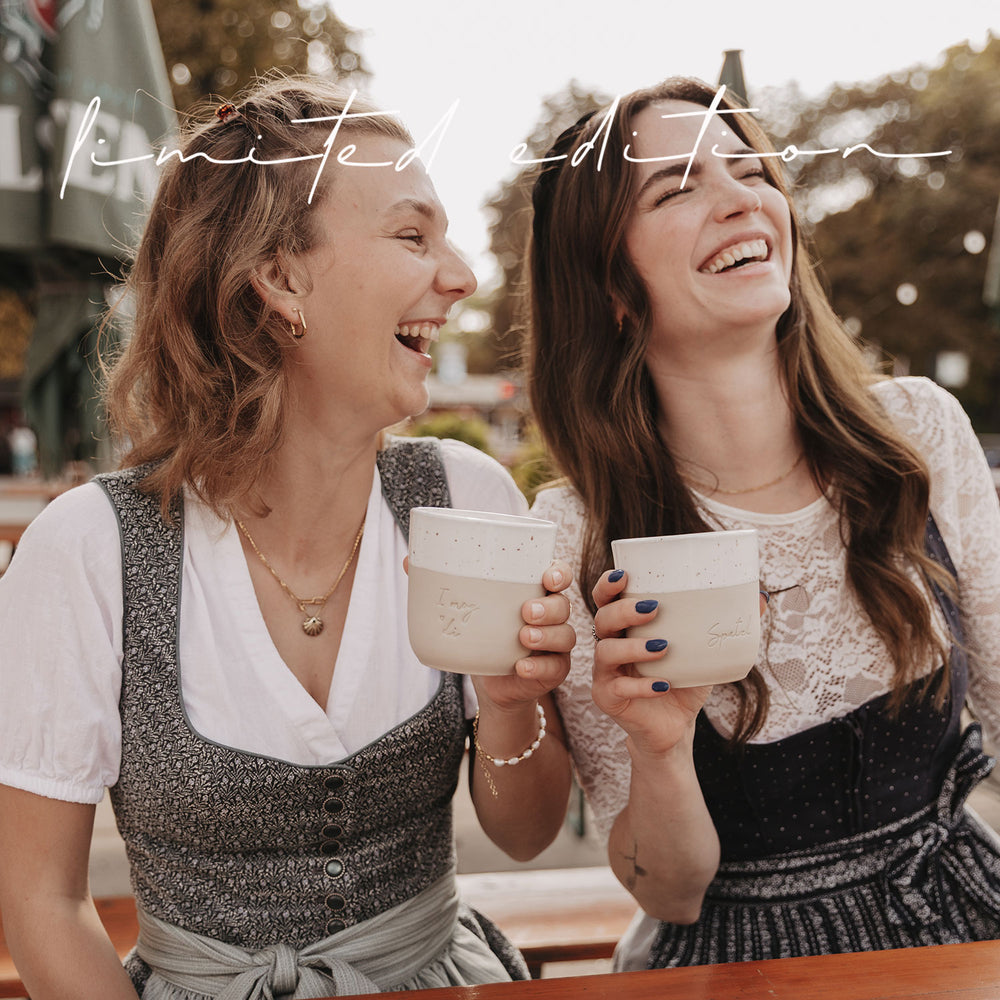 Zwei lachende Frauen im Dirndl halten die limitierten Becher „I mog di“ und „Spatzl“ hoch.