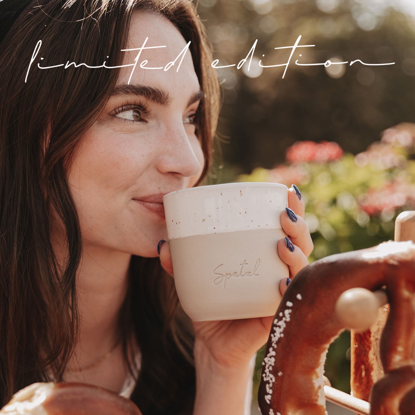 Frau mit blauer Maniküre trinkt aus dem limitierten Becher „Spatzl“ neben einer großen Brezel.