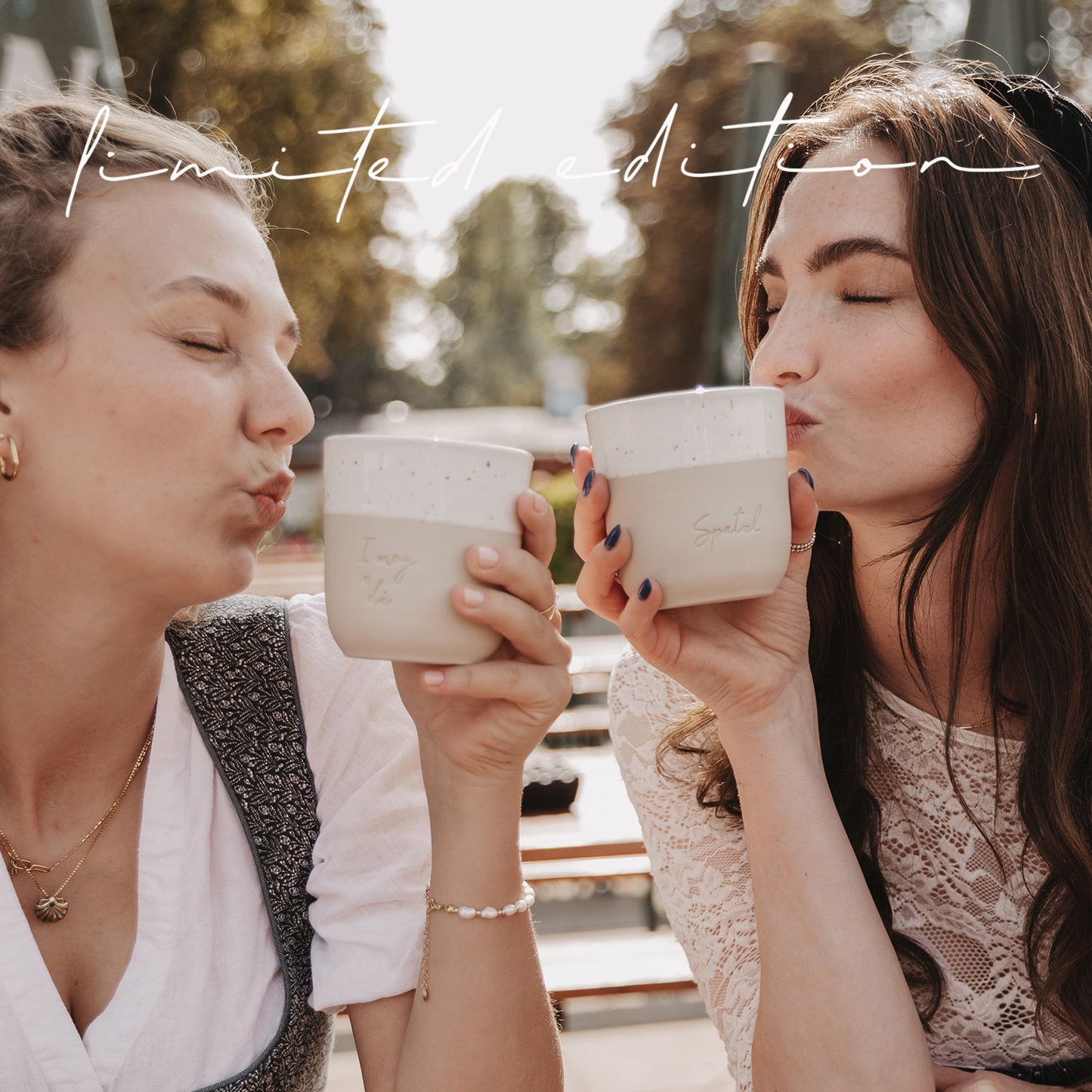 Zwei Frauen halten die Becher „I mog di“ und „Spatzl“ in der Hand und pusten Küsse darauf.