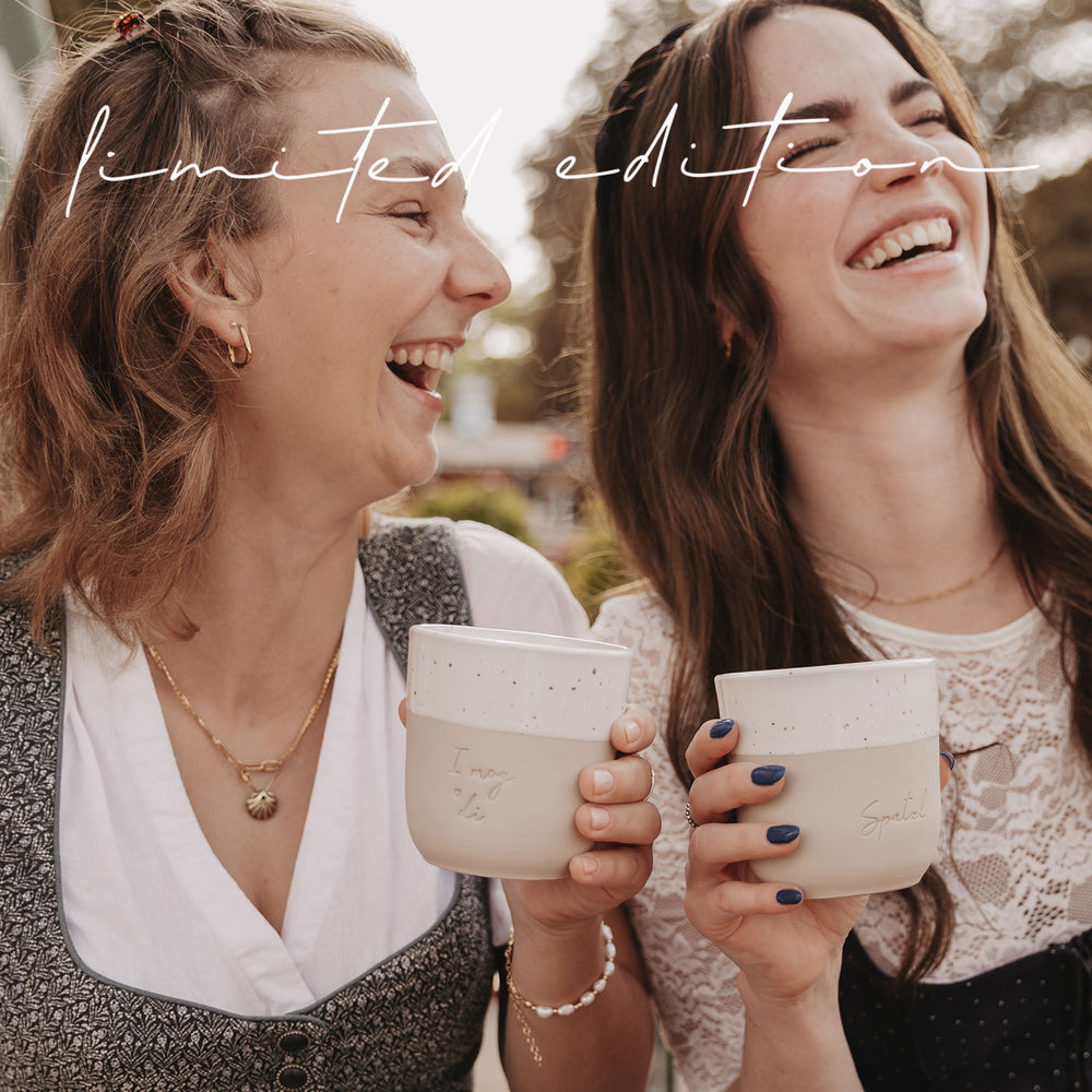 Zwei Freundinnen lachen herzlich mit den limitierten Bechern „I mog di“ und „Spatzl“ in der Hand.