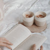 Becher „Me Time“ und „Kleine Auszeit“ mit Milchkaffee auf Bett, daneben geöffnetes Buch – Limited Edition.