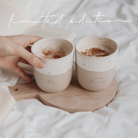 Zwei Keramikbecher Me Time und Kleine Auszeit mit Milchschaum auf Holztablett.