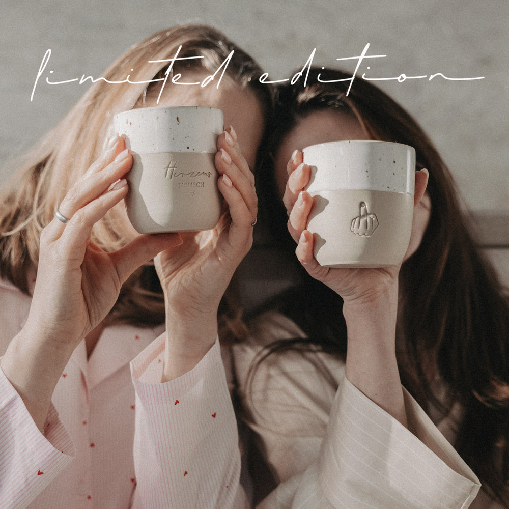 Zwei Frauen halten Herzensmensch- und Mittelfinger-Becher der Limited Edition vor sich, beige-weiß mit Keramikstruktur.