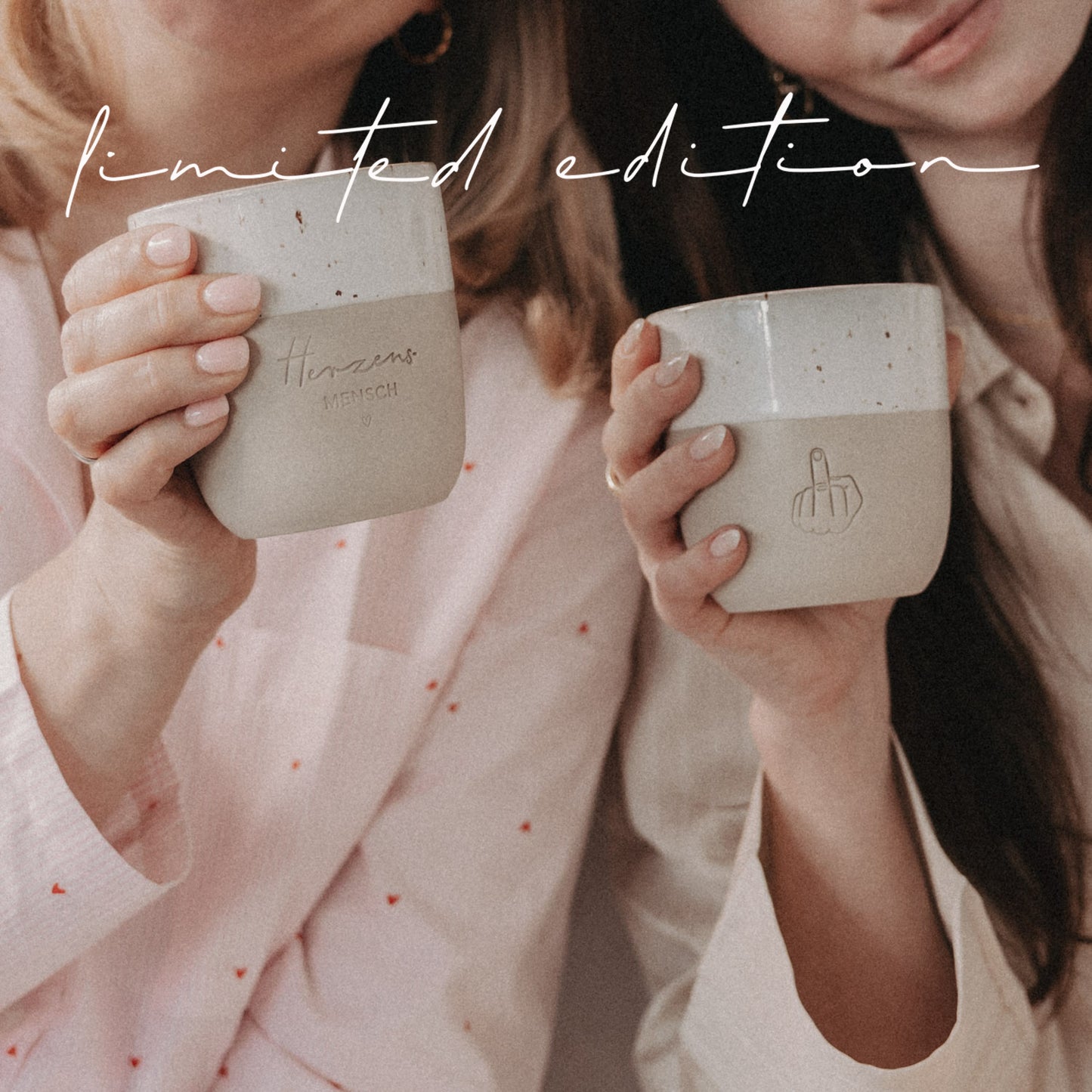 Zwei Frauen halten beige-weiße Limited-Edition-Becher mit Herzensmensch- und Mittelfinger-Motiv und lächeln in die Kamera.