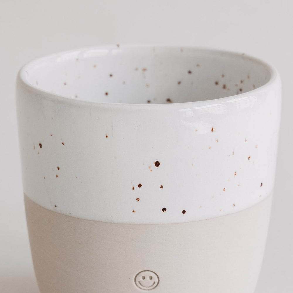 Keramikbecher mit weißer Sprenkelglasur und Smiley-Prägung, Fokus auf die glatte, helle Oberfläche.