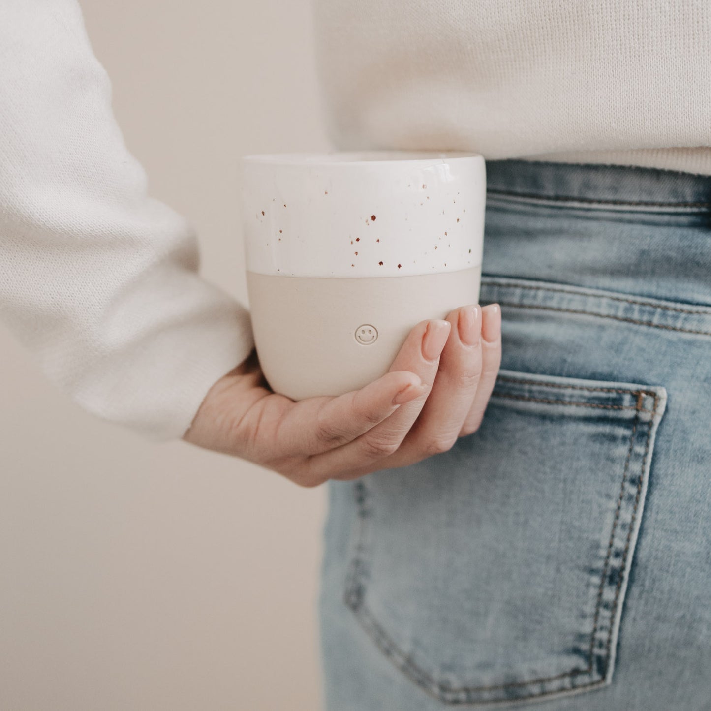 Person hält hellen Keramikbecher mit Smiley-Prägung und weißer Sprenkelglasur locker in der Hand.