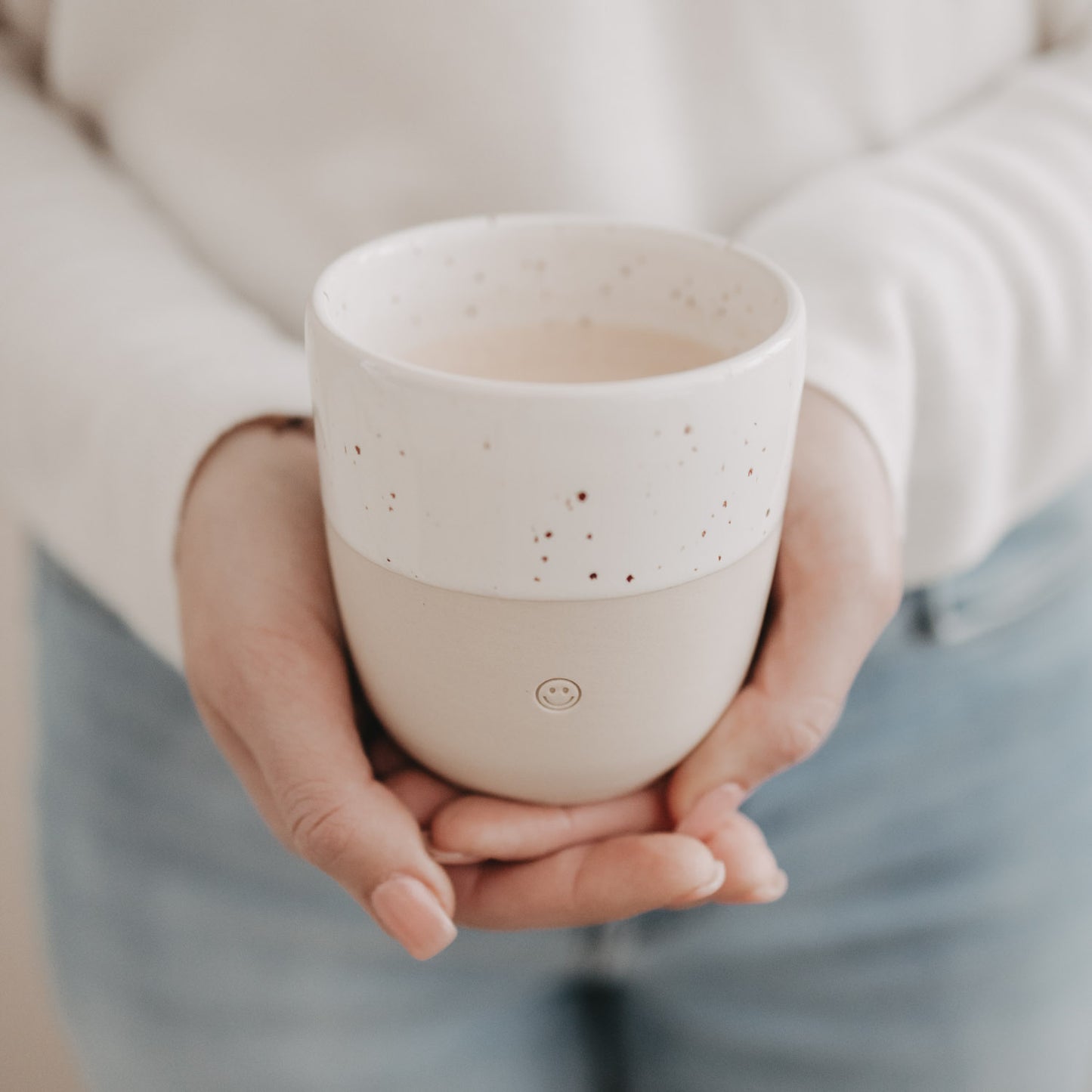 Heller Keramikbecher mit Smiley-Relief, von beiden Händen gehalten, weiße Sprenkelglasur oben.