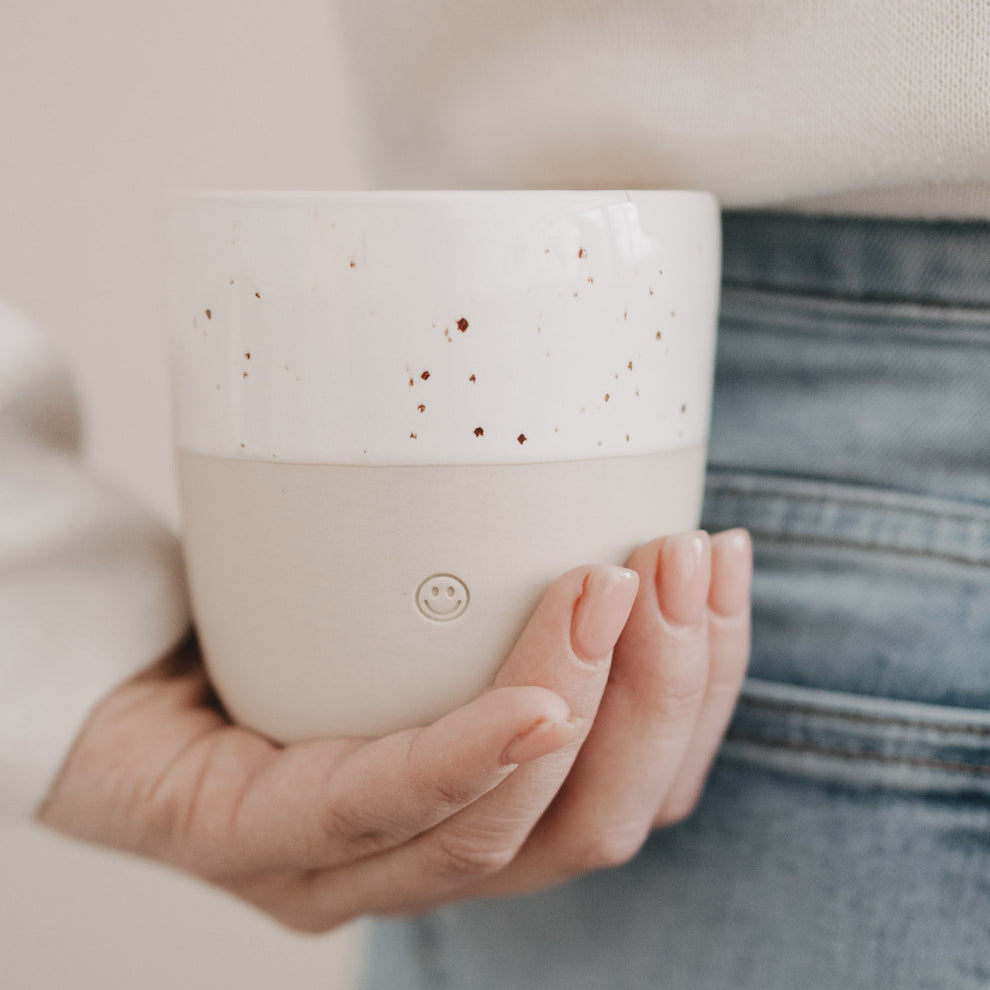 Keramikbecher in Naturtönen mit Smiley-Prägung, Nahaufnahme einer Hand, die den Becher seitlich hält.