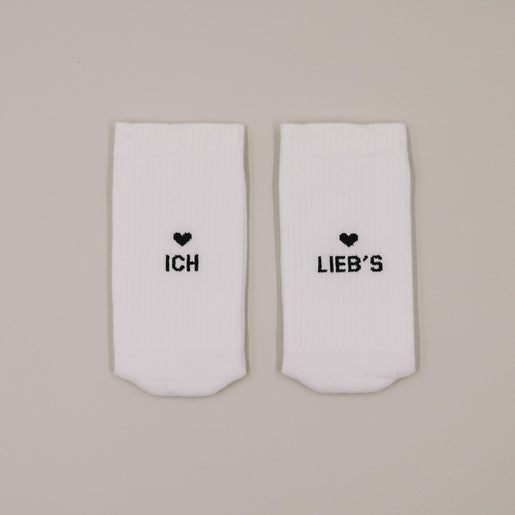 Flachliegende weiße Socken mit Aufdruck „Ich lieb’s“ und kleinem Herzmotiv