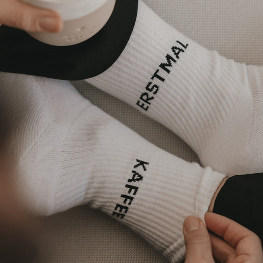 Close-up weißer Socken „Erstmal Kaffee“ mit schwarzer Hose und Kaffeebecher