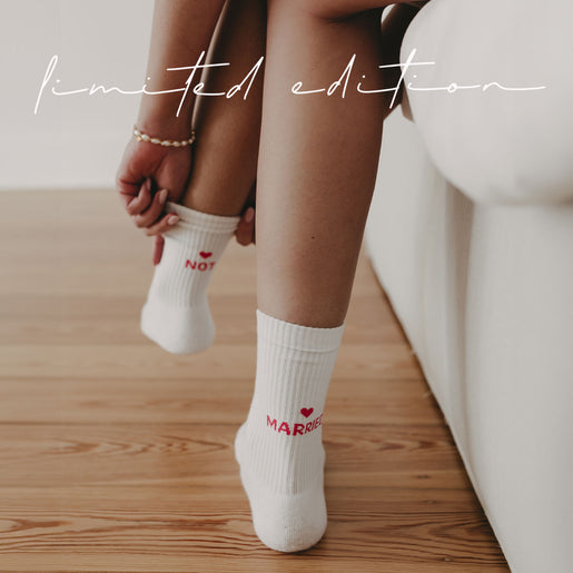 Nahaufnahme: Frau sitzt am Rand eines Sofas auf Holzboden und zieht weiße Socke mit pinker Aufschrift NOT MARRIED hoch. Auf dem Bild steht in Handschrift Limited Edition.