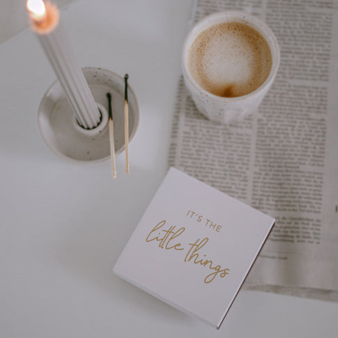 Streichholzschachtel mit goldener Aufschrift „It’s the little things“ neben Kerze und Kaffee auf Zeitung.