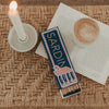 Streichholzschachtel „Sardinen“ in Blau und Rosa mit langen Streichhölzern neben Kerze und Kaffee