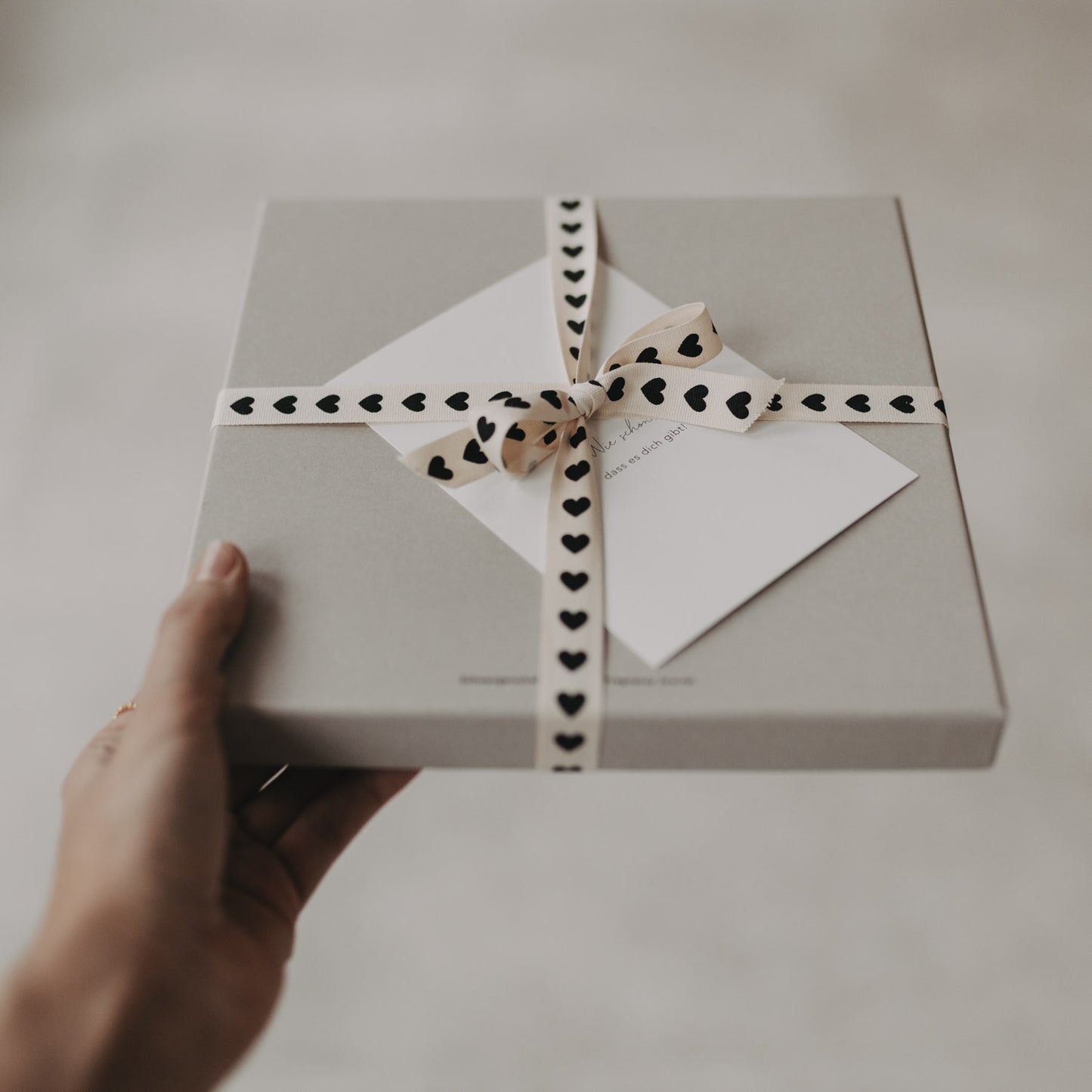 Geschenkbox mit Herz-Geschenkband und Karte, gehalten in der Hand auf hellem Hintergrund.
