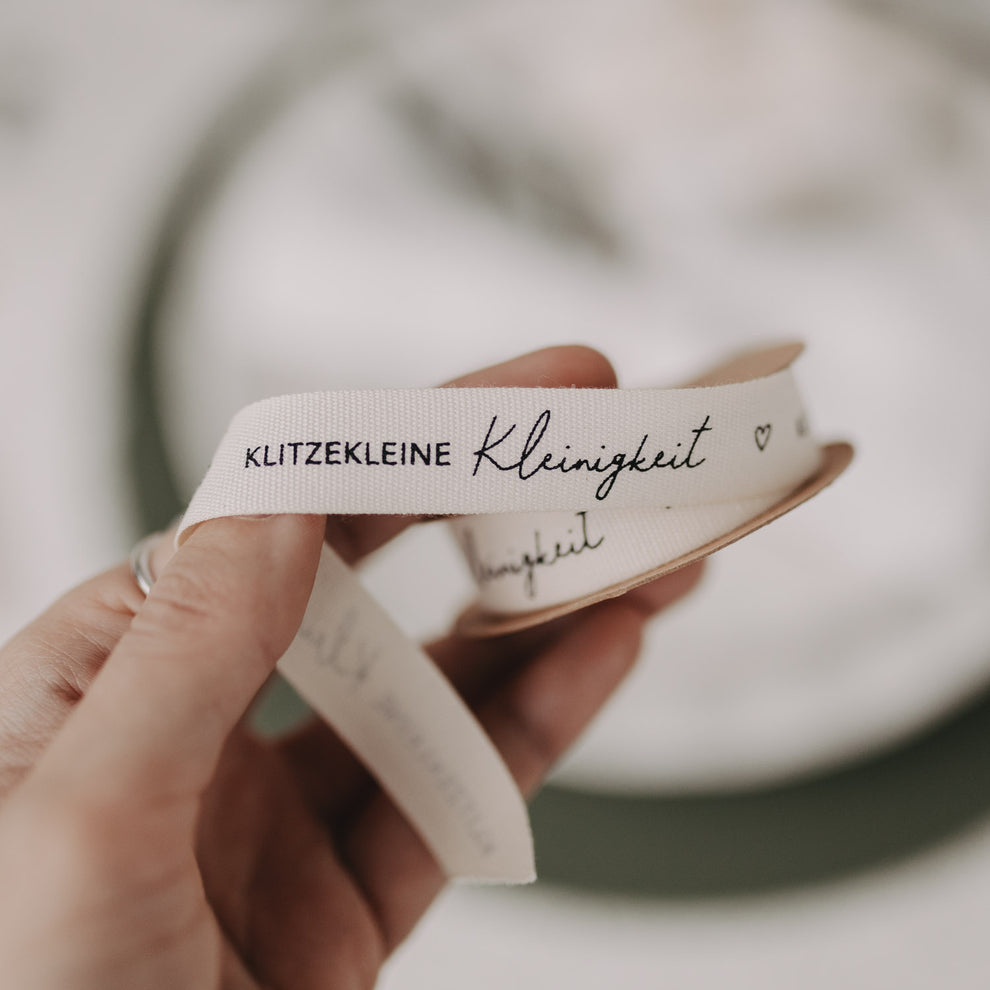 Hand hält weißes Stoff-Geschenkband mit Aufdruck „Klitzekleine Kleinigkeit“.